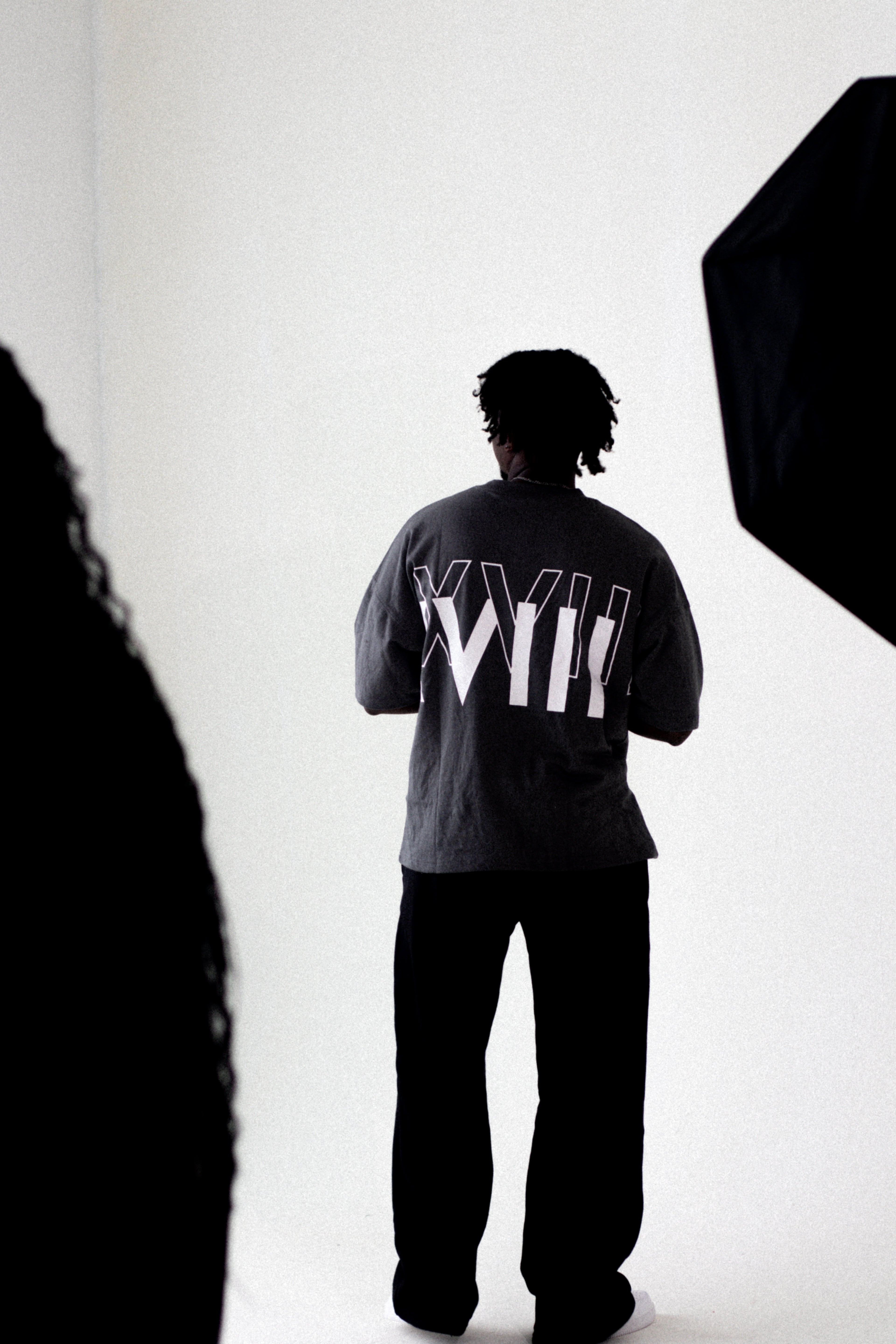 Person wearing a dark gray exactitudeworld t-shirt brand with 'XVII' printed on the back, standing against a white background.